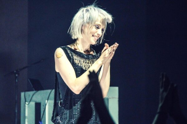 A light-skinned woman with silvery hair puts her hands together on a stage