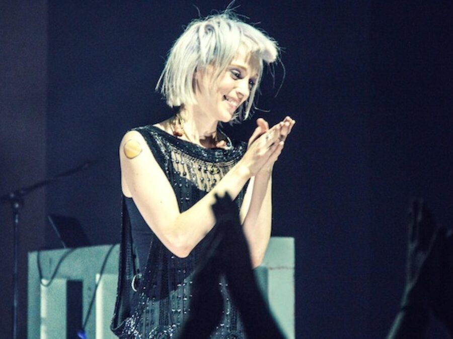 A light-skinned woman with silvery hair puts her hands together on a stage