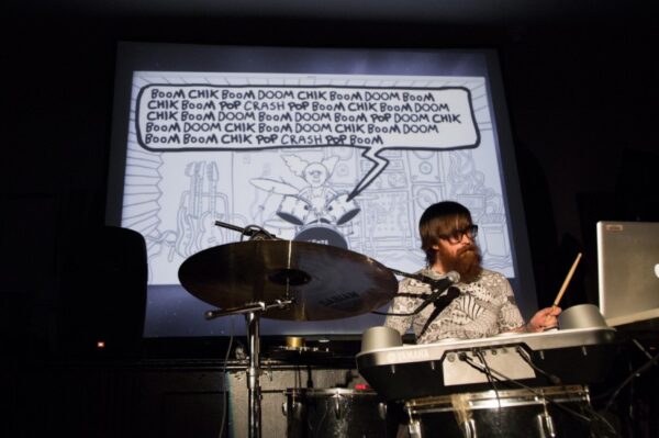 A bearded person with glasses behind a drum kit with a microphone and computer. On the screen behind them is a word blurb projected on the screen filled with words that describe the sounds of a drum.