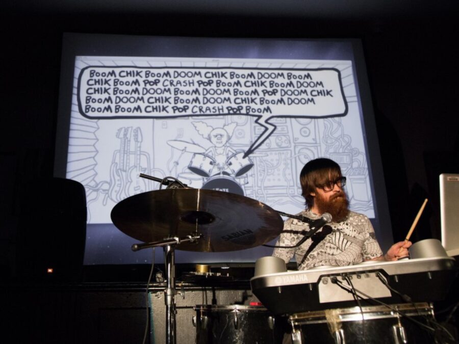 A bearded person with glasses behind a drum kit with a microphone and computer. On the screen behind them is a word blurb projected on the screen filled with words that describe the sounds of a drum.