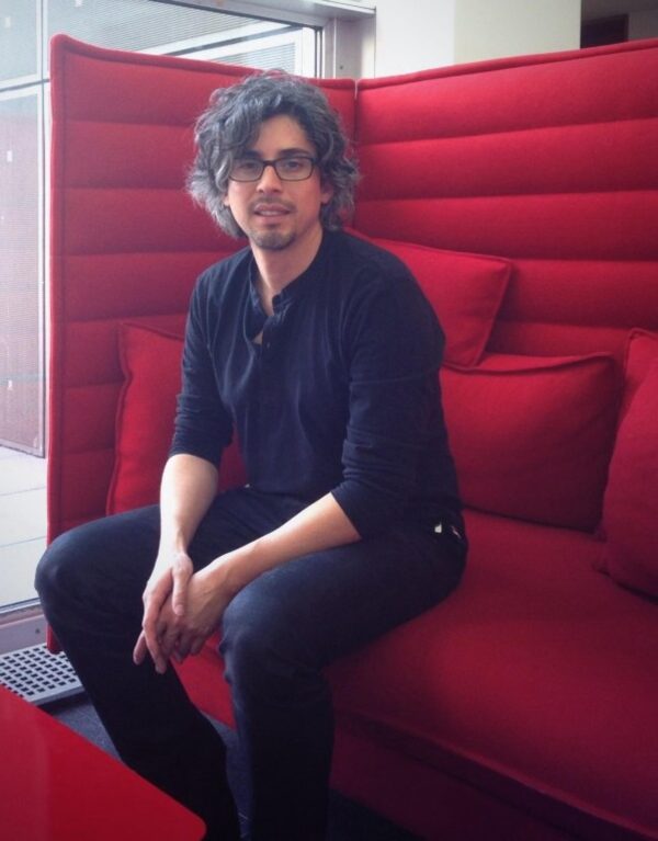 A male with light skin and dark curly hair dressed in black sits on a high-backed red couch
