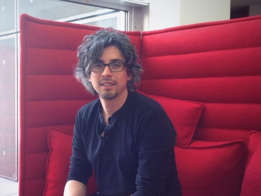 A male with light skin and dark curly hair dressed in black sits on a high-backed red couch