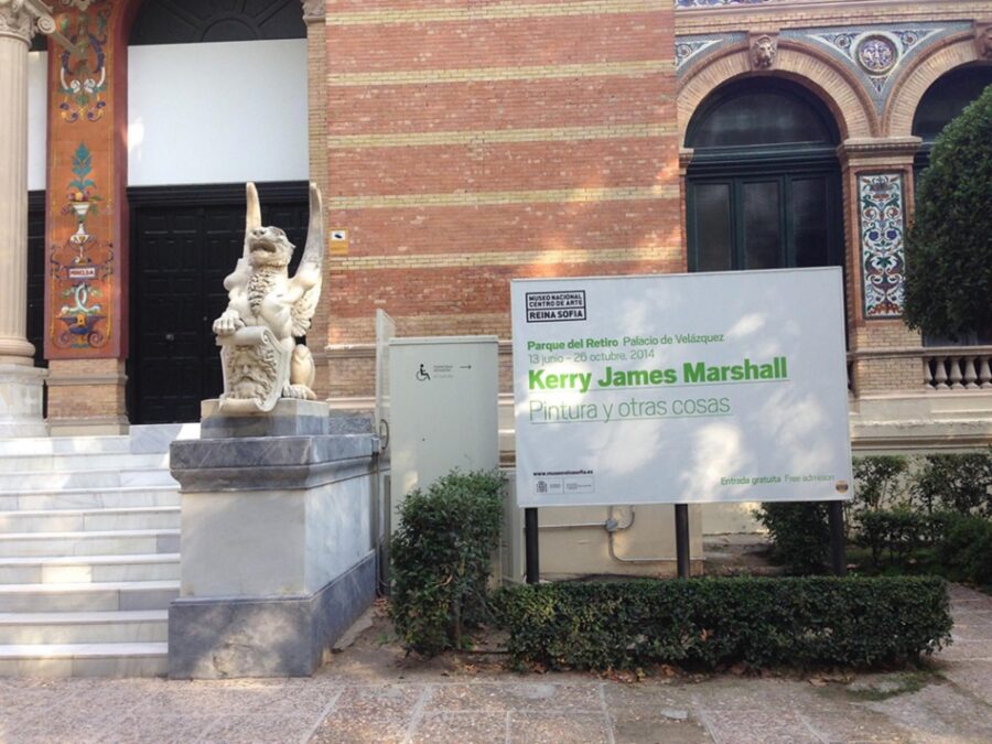 A large plain white sign reading Kerry James Marshall is posted next to a gargoyle that guards the steps up to a building with intricate brickwork