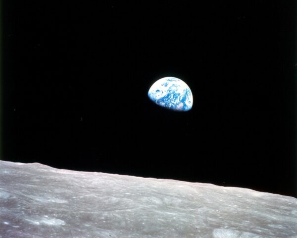 A vibrant blue-and-white Earth peeking over the barren, cratered grey lunar horizon into the, pitch-black void of space.