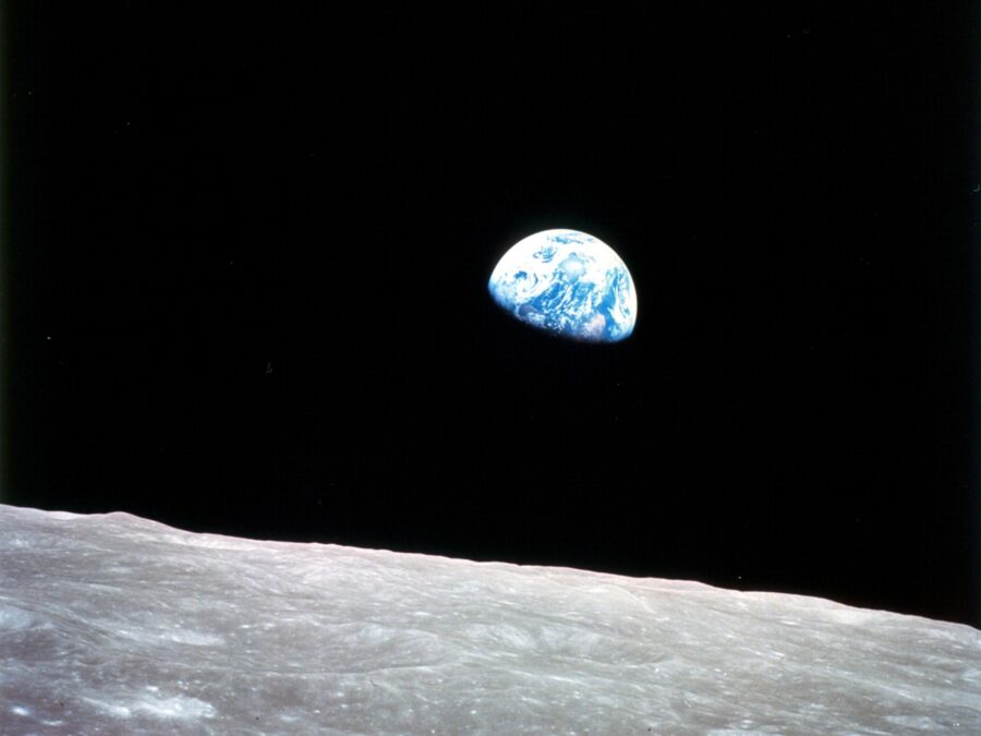 A vibrant blue-and-white Earth peeking over the barren, cratered grey lunar horizon into the, pitch-black void of space.