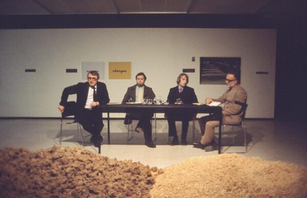 Four men sit at a table in a gallery space. In front of them are two piles of grain or sand