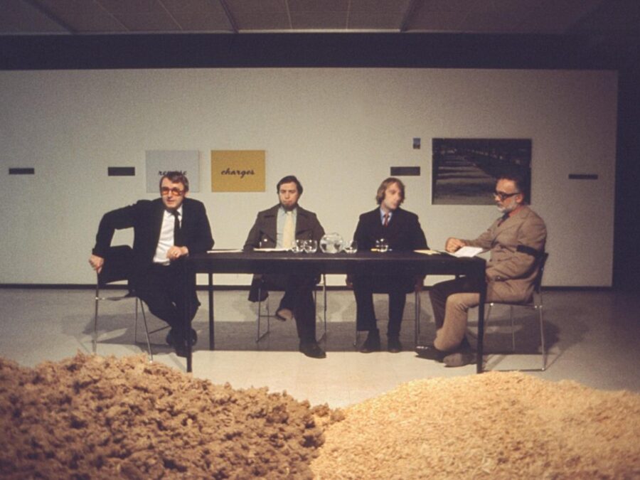 Four men sit at a table in a gallery space. In front of them are two piles of grain or sand