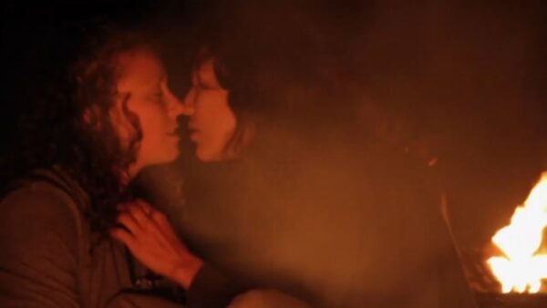 Two people lit red by the fire that is just visible in the bottom right. One leans in for a kiss, both of their lips parted.