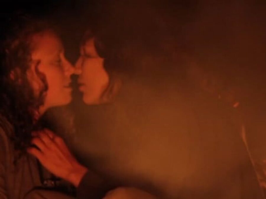 Two people lit red by the fire that is just visible in the bottom right. One leans in for a kiss, both of their lips parted.