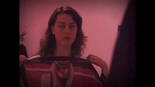 A person holds a sweater up to their upper torso in a red-tinted video still where their eyes have been caught mid-blink