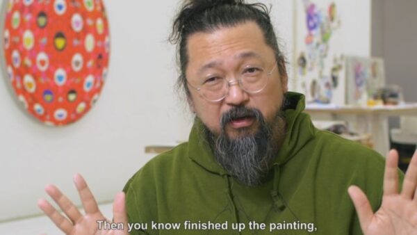 Takashi Murakami, a Japanese man sporting a greying beard and small wire-frame glasses, gestures with both hands while sitting in his studio, some of his artwork just visible on the walls and table behind him.