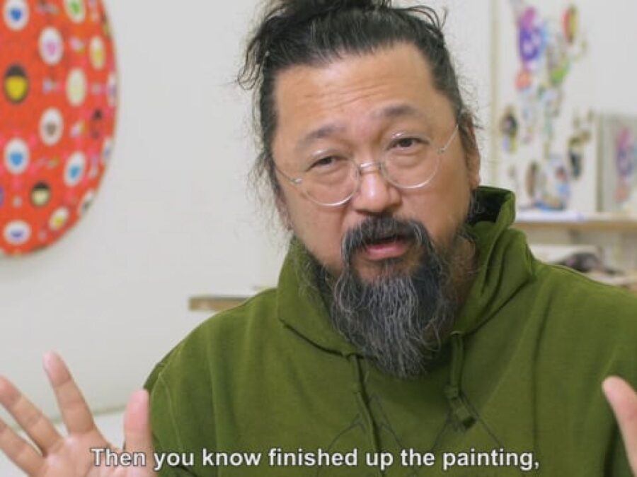 Takashi Murakami, a Japanese man sporting a greying beard and small wire-frame glasses, gestures with both hands while sitting in his studio, some of his artwork just visible on the walls and table behind him.