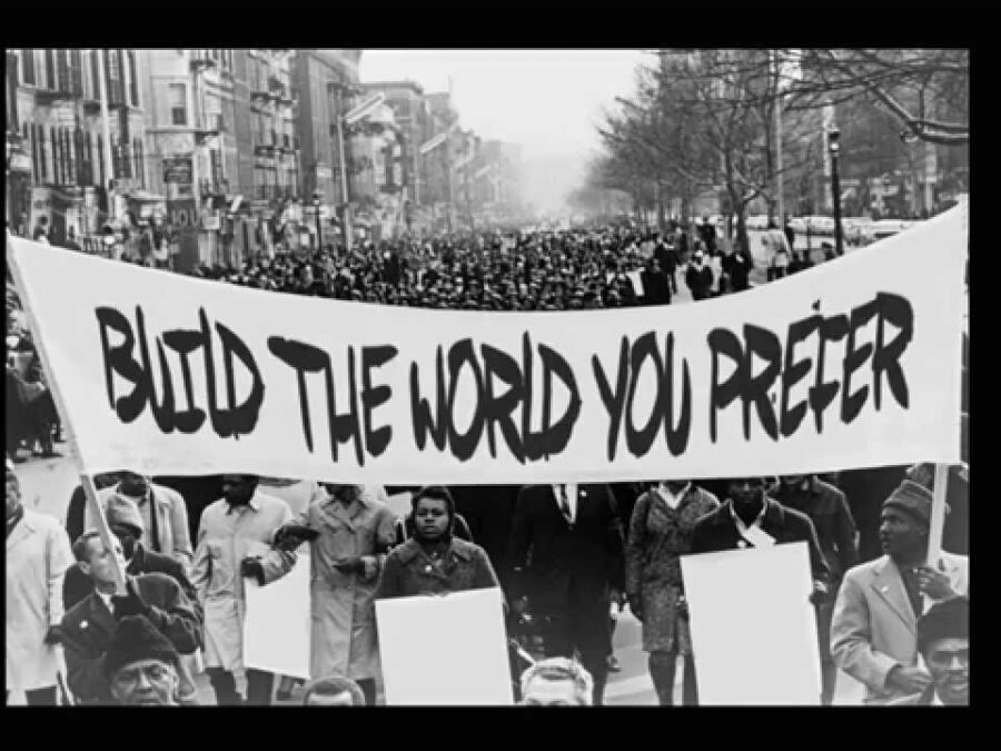 Black and white photo of a crowd of people holding a banner stating Build the world you prefer
