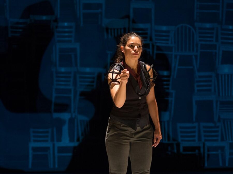 A singular, spotlit performer in all black points toward the audience in front of a background of stacked chairs
