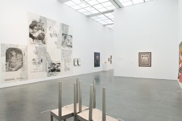 A white-walled gallery space filled with various artworks, including a metal sculpture on the foreground. The space extends to another room just out of view.