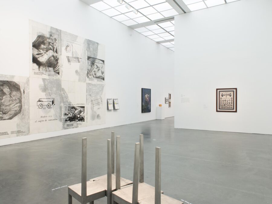 A white-walled gallery space filled with various artworks, including a metal sculpture on the foreground. The space extends to another room just out of view.