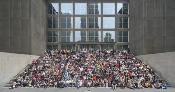 A photograph, captured from a distance, depicts a large group of people seated on the MCA's front steps.