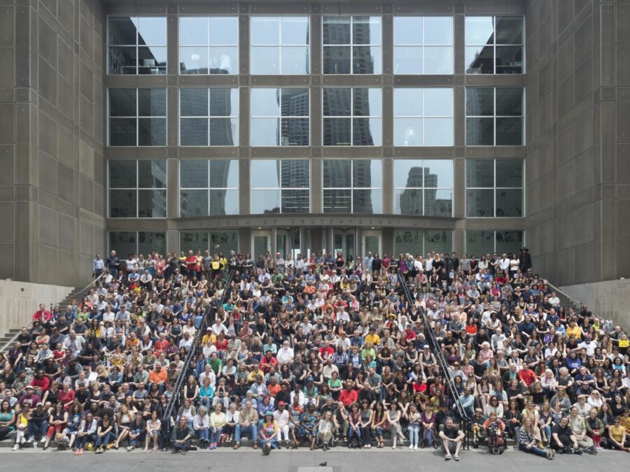 A photograph, captured from a distance, depicts a large group of people seated on the MCA's front steps.