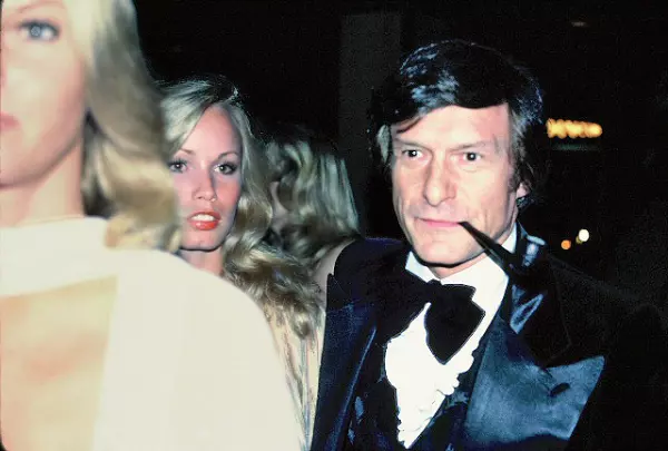 A young Hugh Hefner in a tux with a pipe hanging out of his mouth standing behind and next two two blondes that are barely visible