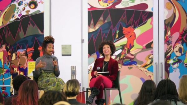 Two people sit in front of a colorful wall holding microphones as a group of people look on