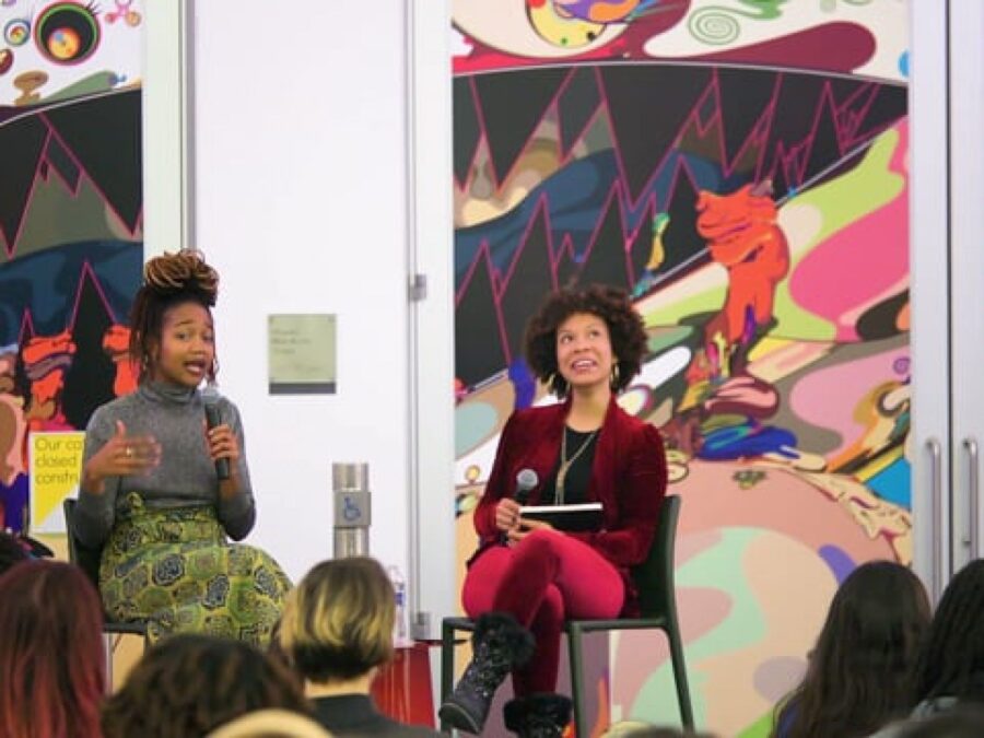 Two people sit in front of a colorful wall holding microphones as a group of people look on