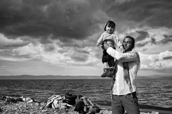 A tan-skinned man holds up a young, smiling child in front of a wide expanse of choppy water, distant low-lying hills, and a cloudy sky.