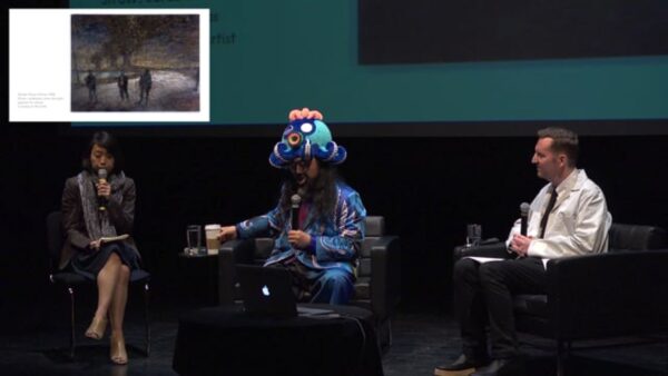 Three people sit on a stage with a powerpoint presentation behind them and as a picture-in-picture in the upper left corner. The middle person wears a colorful cartoon-like octopus hat.
