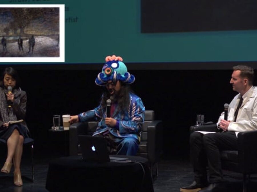 Three people sit on a stage with a powerpoint presentation behind them and as a picture-in-picture in the upper left corner. The middle person wears a colorful cartoon-like octopus hat.