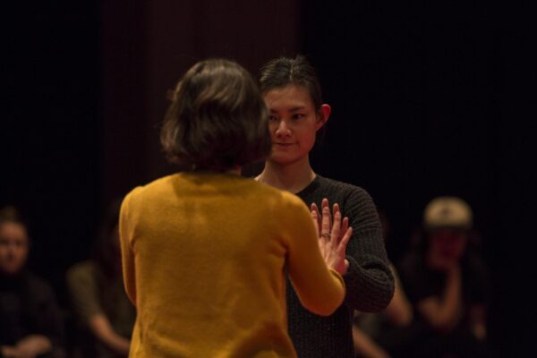In a dimly lit room two people stand close together palms touching as other people behind them look on
