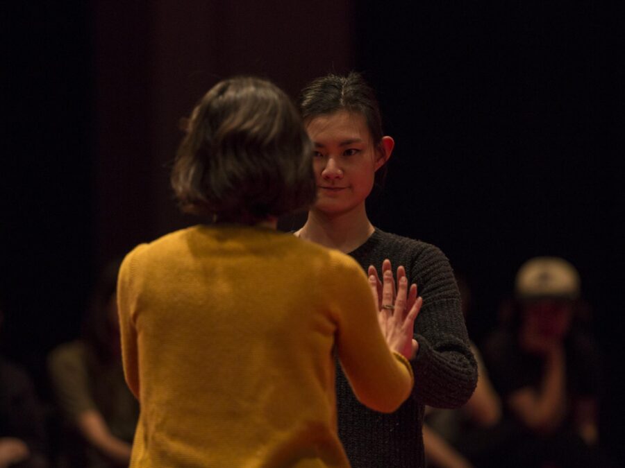In a dimly lit room two people stand close together palms touching as other people behind them look on