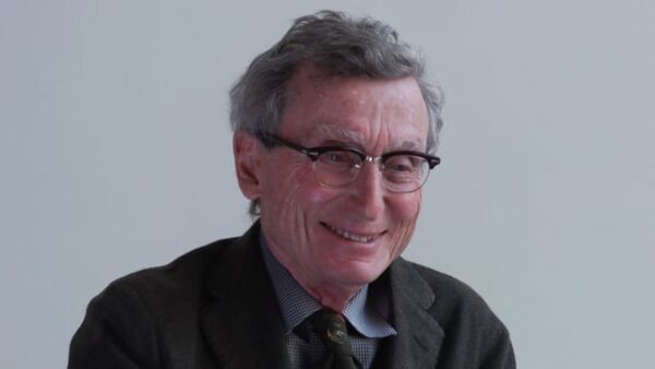 An older man in glasses smiling against a gray backdrop