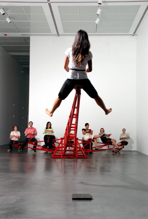 One person is raised up by an uneven red teeter-totter in which six people sit on the other side.