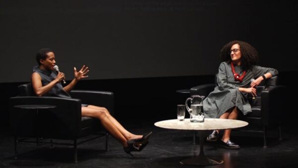 Two people sit in black chairs on a black stage with a white table between them