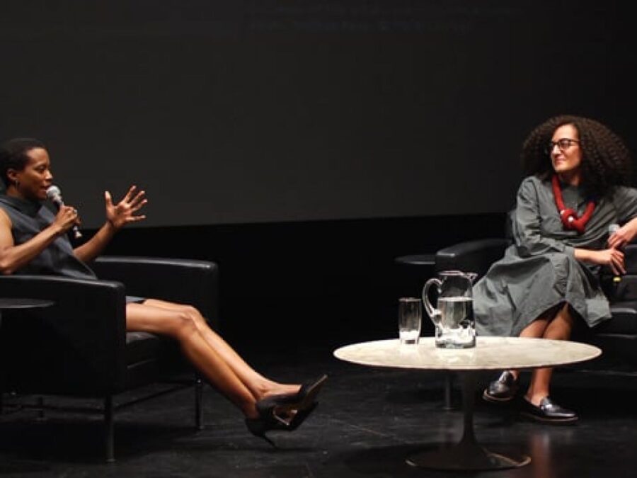 Two people sit in black chairs on a black stage with a white table between them