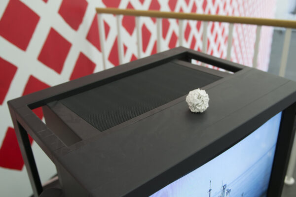 A white crumpled piece of paper on a tv in front of a white railing and wall filled with red diamonds