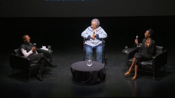 Three women sit on a black stage holding microphones