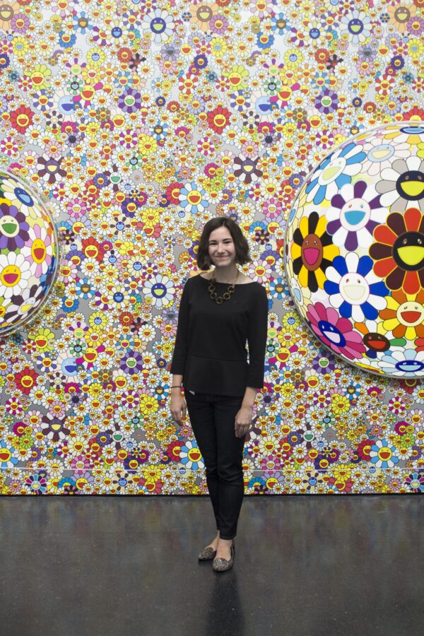 A person poses between two flower orbs in front of a wall covered in happy daisy faces