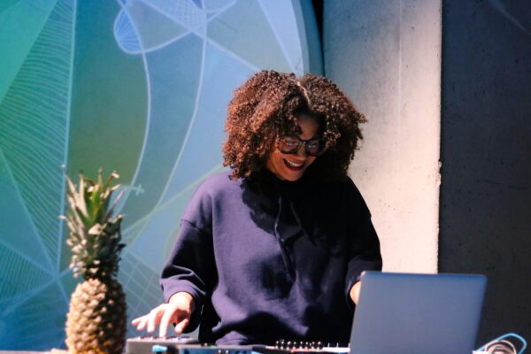 A female-presenting person with full brown curly hair smiles down at her laptop while adjusting sound equipment. A pineapple is placed at her right.