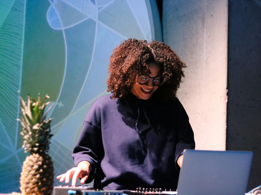 A female-presenting person with full brown curly hair smiles down at her laptop while adjusting sound equipment. A pineapple is placed at her right.