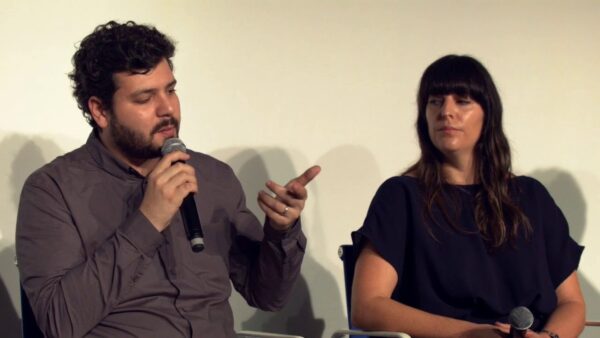 Two people sit in a yellow-tinged room. The person on the left gestures with their hands while holding a microphone