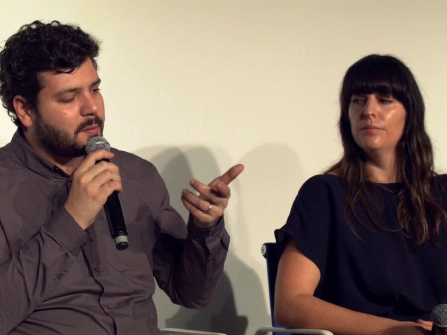 Two people sit in a yellow-tinged room. The person on the left gestures with their hands while holding a microphone