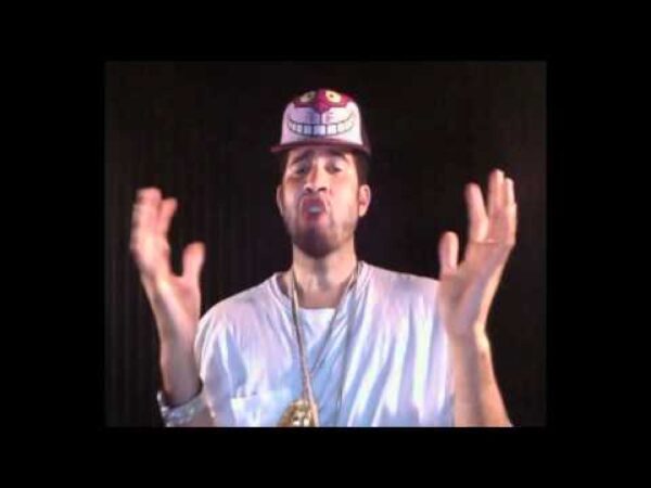 A male presenting person with light brown skin wearing a Cheshire Cat hat and large gold-chained necklaces gestures with his hands against a black backdrop