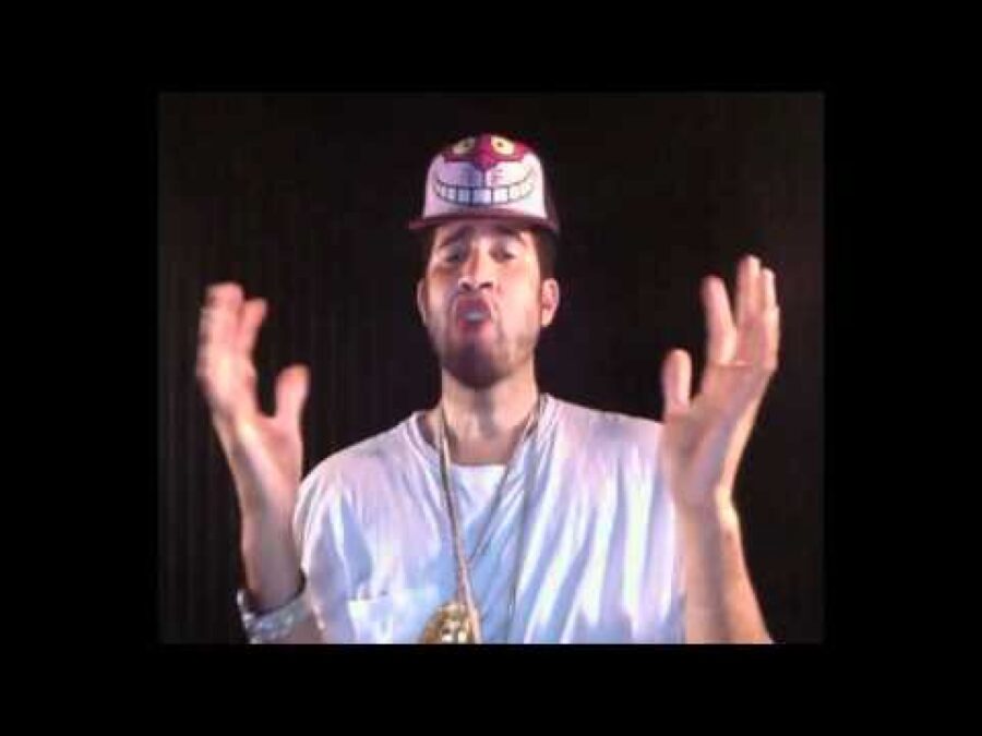 A male presenting person with light brown skin wearing a Cheshire Cat hat and large gold-chained necklaces gestures with his hands against a black backdrop