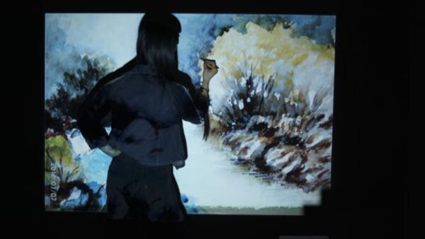 In a dark room, a person with long dark hair faces a large canvas and holds a tool up to it with their right hand