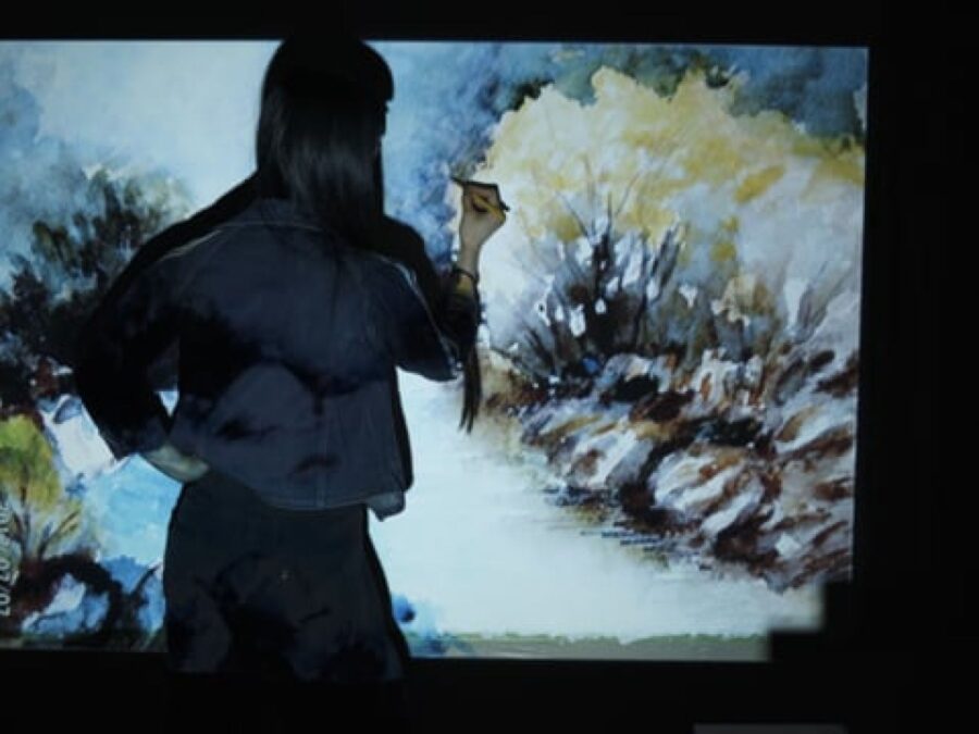 In a dark room, a person with long dark hair faces a large canvas and holds a tool up to it with their right hand