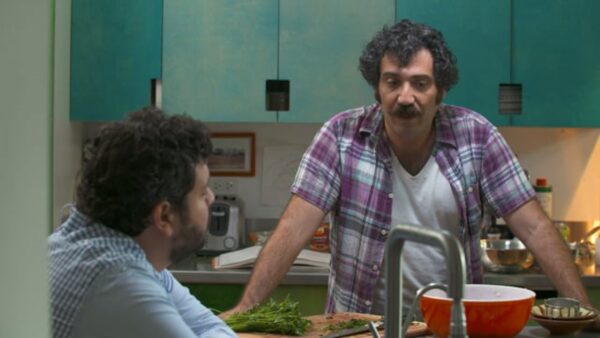 Michael Rakowitz, a man with dark curly hair and a mustache, leans against a kitchen counter, talking to a person sitting