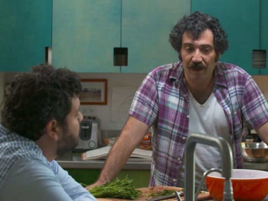 Michael Rakowitz, a man with dark curly hair and a mustache, leans against a kitchen counter, talking to a person sitting