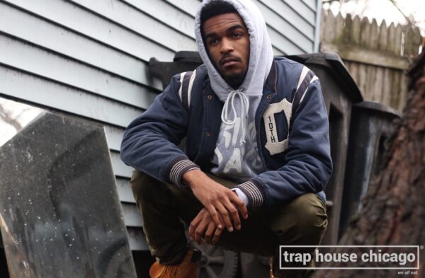 A model with light brown skin squats down next to a garbage bin outside. He is wearing a grey hoodie with the words Crime Pays barely legible from underneath a letterman jacket. The words trap house Chicago appear in white font within a box in the bottom-right corner of the image