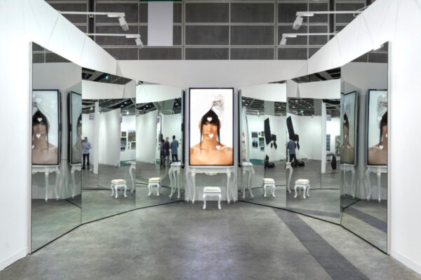 A screen with a three-quarter image of a person in it rests atop a white vanity with a matching chair in front of it. A wall of mirrors appears on either side of this, reflecting the set of objects as well as the gallery space it is in.
