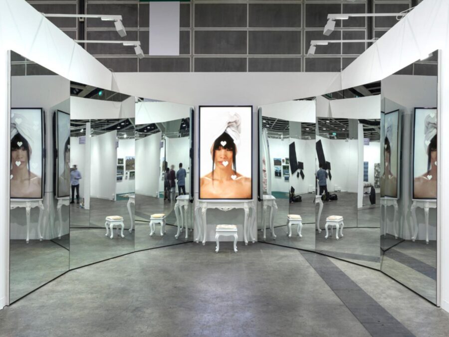 A screen with a three-quarter image of a person in it rests atop a white vanity with a matching chair in front of it. A wall of mirrors appears on either side of this, reflecting the set of objects as well as the gallery space it is in.